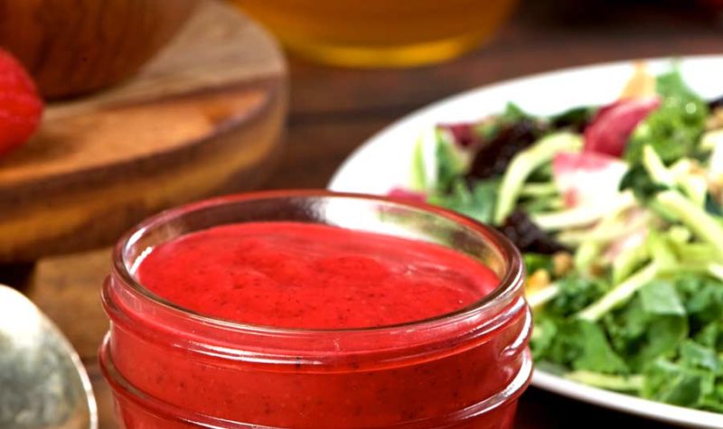 A small glass jar filled with raspberry vinaigrette with a spoon and side salad.