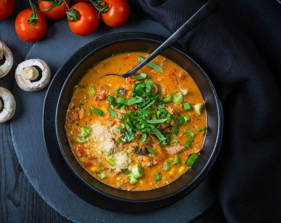 A black bowl filled with pizza soup with a spoon inside, and fresh mushrooms and tomatoes on the side.