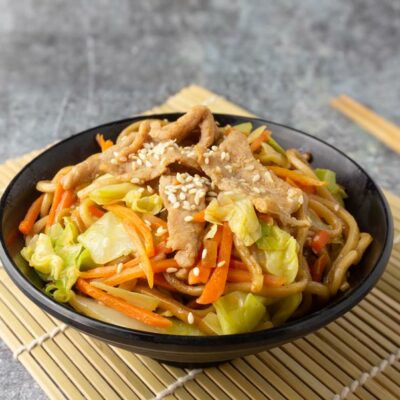 A black bowl filled with noodles and asian veggies, and pork.