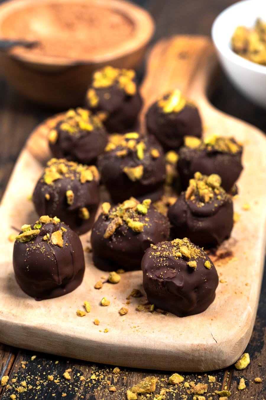 A cutting board with chocolate truffles on top, covered with pieces of pistachios.