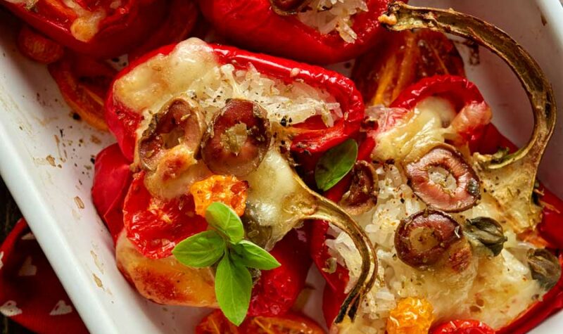 Red bell peppers stuffed vegetables and mozzarella cheese seasoned with herbs in a baking dish on a black wooden table
