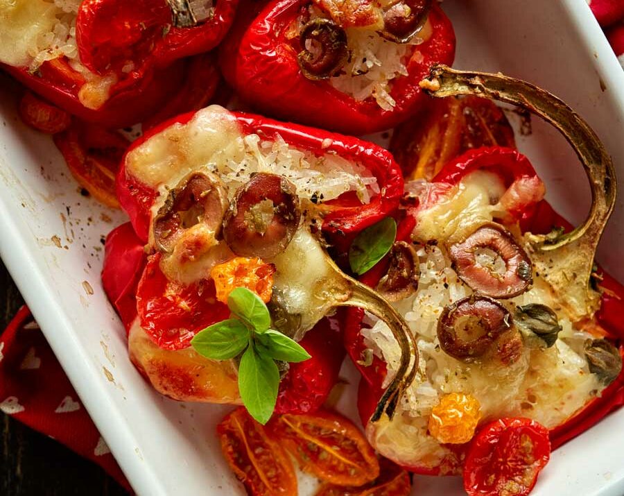 Red bell peppers stuffed vegetables and mozzarella cheese seasoned with herbs in a baking dish on a black wooden table