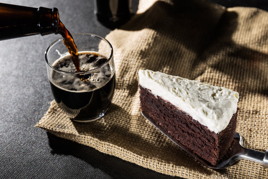 A piece of chocolate cake with beer being poured in the background.